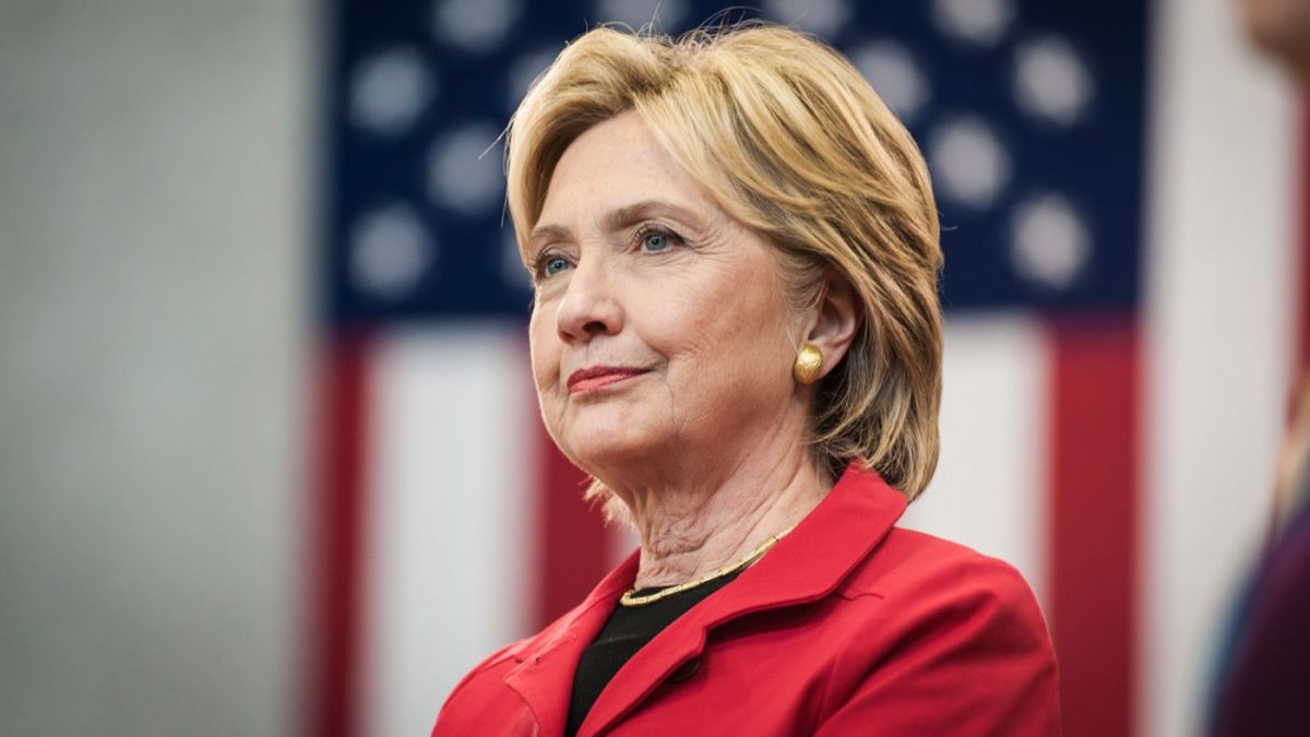 Hillary Clinton looks meditative and proud accompanied by the flag of The United States of America. Image Credit: El Ágora
